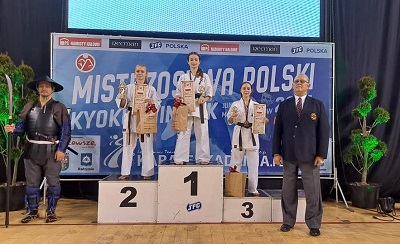Patrycja stojąca na podium, obok niej dziewczyny, które zajęły pierwsze i drugie miejsce. Wszystkie ubrane w kimona trzymają dyplomy i puchary.